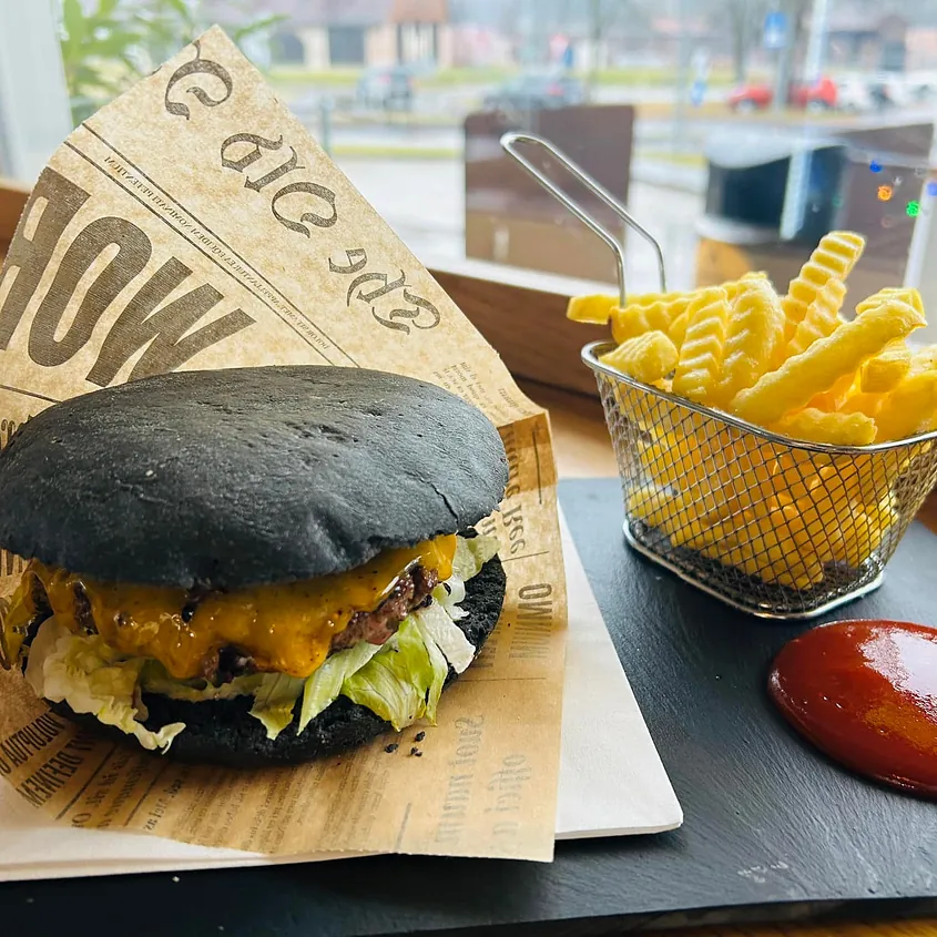 Grill & Chill Black Bun Burger mit Cheddar und Salat, serviert mit Pommes im Metallkörbchen und Ketchup auf Schieferplatte