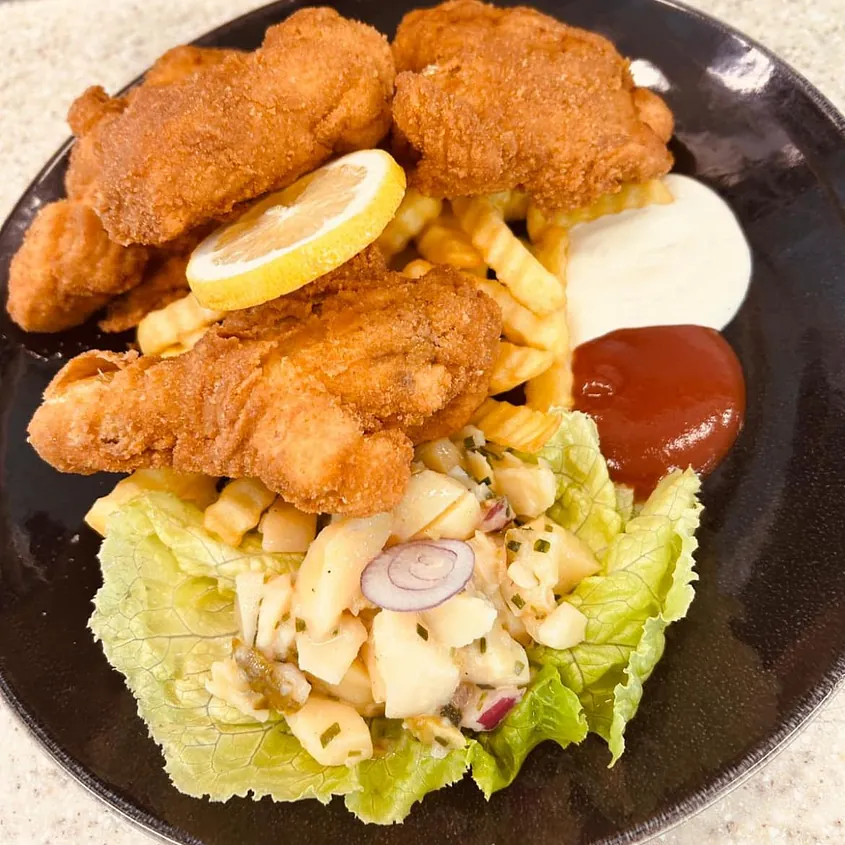 Goldbraun paniertes Backhendl mit knusprigen Pommes, hausgemachtem Kartoffelsalat und Dips auf schwarzem Teller